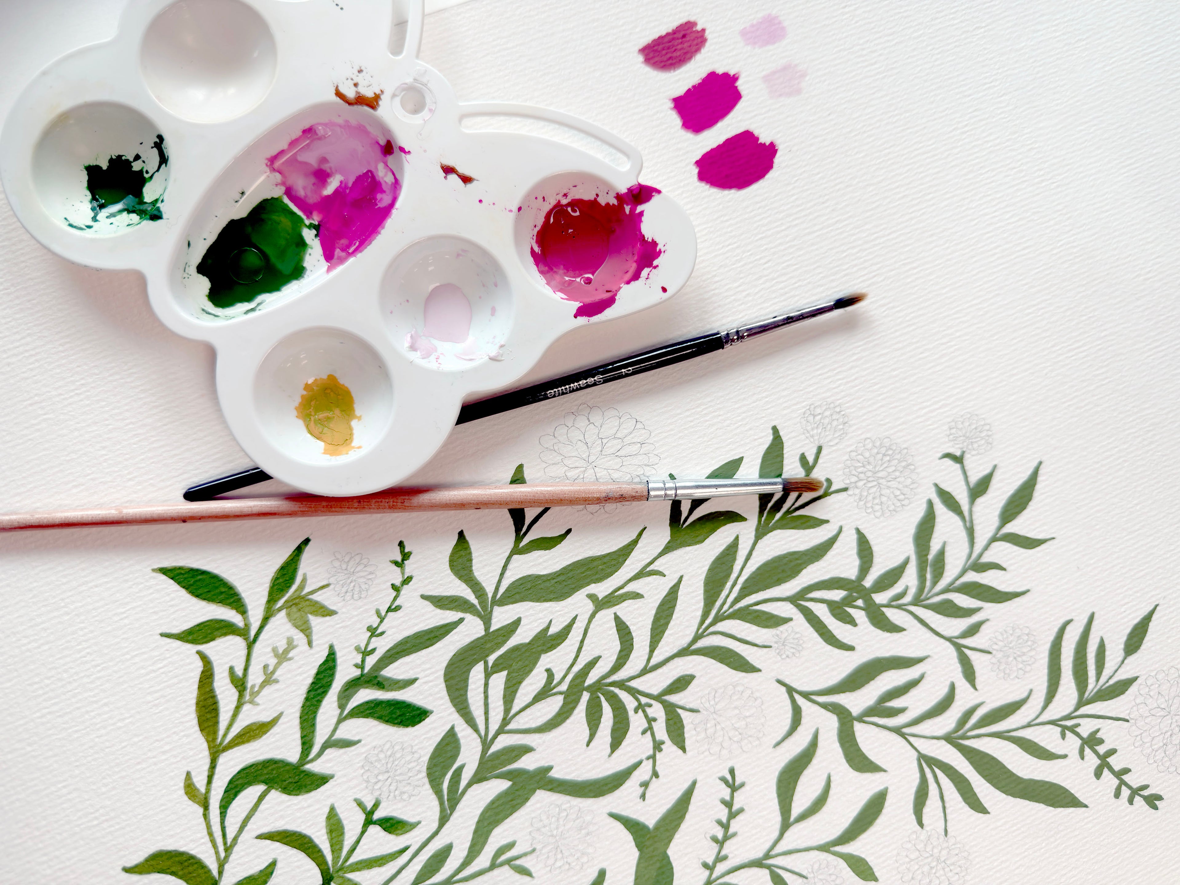 A white paint palette with mixed paint colors and two paintbrushes arranged on a white textured surface, with green botanical leaf illustrations painted across the lower half.